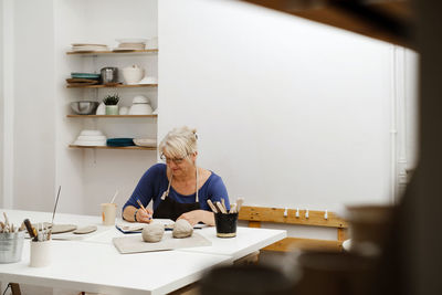 Woman potter planning work at workshop, writing in notebook in light studio her creations. clay