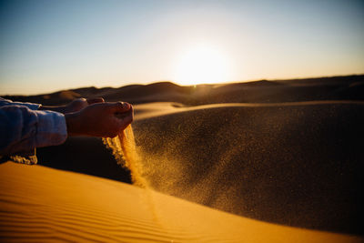 Cropped image of hand pouring sand at desert against sky