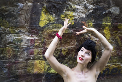 Smiling woman with arms raised standing against stone wall