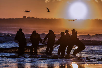 Fishermen pulling net from sea against sky during sunset