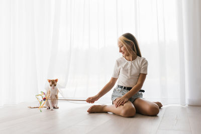 Caucasian teen girl sitting on the floor playing with a cat cornish rex with a stick teaser 