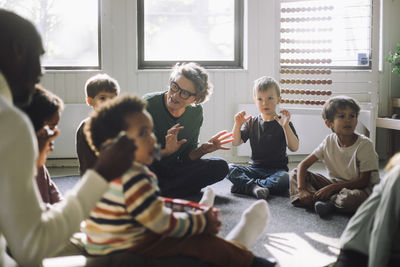 Senior female teacher playing with group of preschool children while sitting in classroom at kindergarten