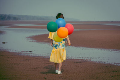 Rear view full length of young woman holding colorful balloons at beach