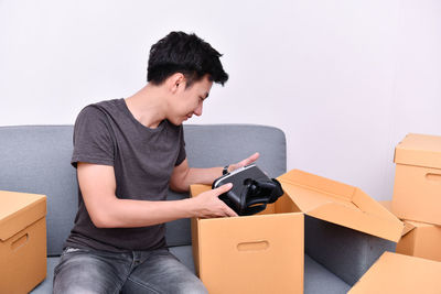 Young man unpacking cardboard boxes sitting on sofa against wall at home