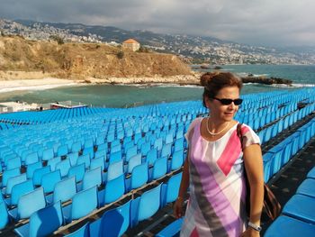 Woman wearing sunglasses while standing by chairs against sea and sky