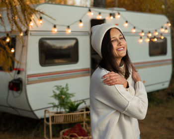 A pensive caucasian woman in a white knitted sweater and a hat is resting near a motorhome in a warm