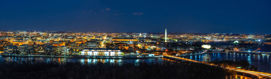 Illuminated buildings by river against blue sky at night