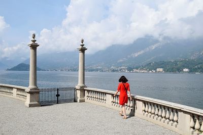 Full length rear view of woman in red dress walking by river against cloudy sky