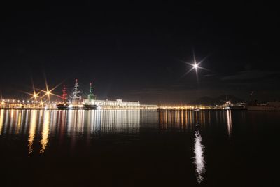 Illuminated city by sea against sky at night