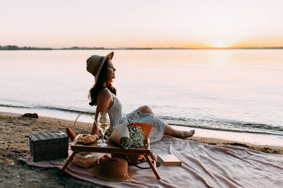 Woman sitting on beach by sea against sky during sunset