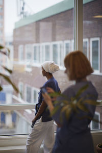 Female doctor standing at hospital corridor and looking through window, another female doctor using cell phone