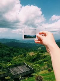 Cropped hand holding grass in field against cloudy sky