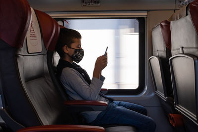 Young boy traveling by train and looking a smart phone. he wears a protective face mask. 