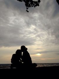 Silhouette couple sitting on beach against sky during sunset