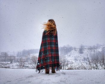 Woman wrapped in blanket standing on snow covered landscape against sky