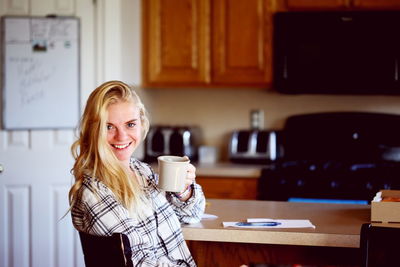 Portrait of smiling young woman having coffee by table in kitchen at home