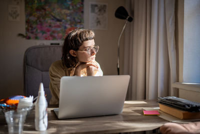 Woman freelancer procrastinating looking at window sitting at desk trying to work on laptop.