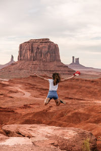 Rear view of woman jumping on rock formation against sky