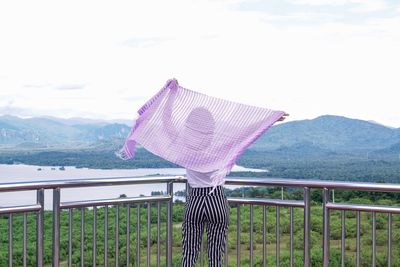 Rear view of woman standing with scarf by railing against sky