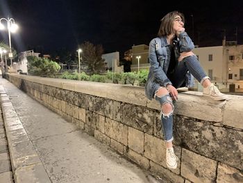 Full length of woman sitting on wall against building at night