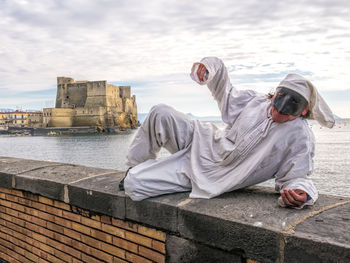 Portrait of man wearing mask lying on retaining wall against sea
