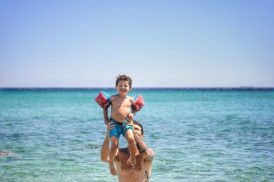 Shirtless father carrying happy son in sea against clear sky