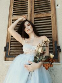 Young woman holding wilted flowers while standing against window outside house