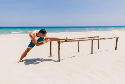 Man exercising on parallel bars at beach against blue sky