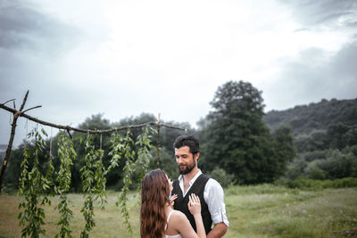 Smiling man looking woman while standing on field against cloudy sky