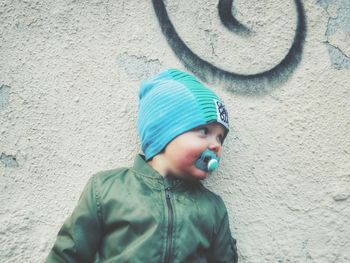 Baby boy with pacifier in mouth standing against wall