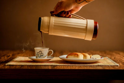 Cropped hand of woman pouring hot tea in cup from container at home