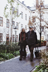 Happy senior couple walking on footpath with luggage outside hotel