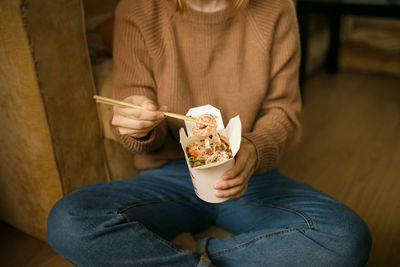 Girl in casual clothes eating chinese noodles from a box sitting on the floor, food delivery