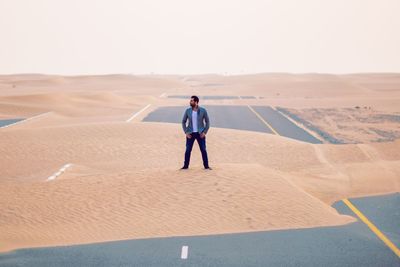 Full length of handsome young man standing on sand at desert against clear sky