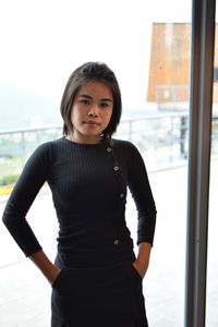 Portrait of young woman standing against window in room