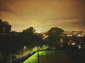 Trees by illuminated city against sky at sunset