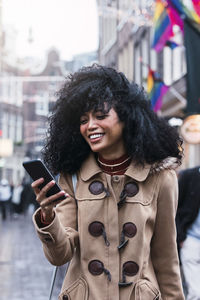 Happy young woman with tousled hair surfing net through smart phone in city