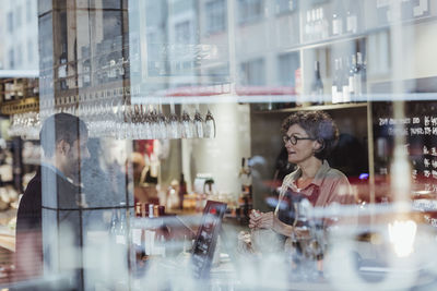 Male customer collecting order from female owner seen through glass in deli store