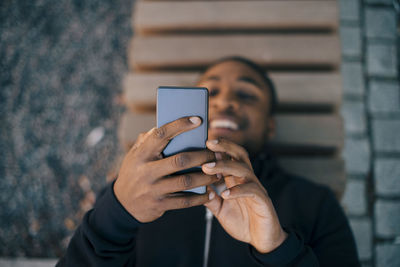 High angle view of smiling man using smart phone while lying on bench