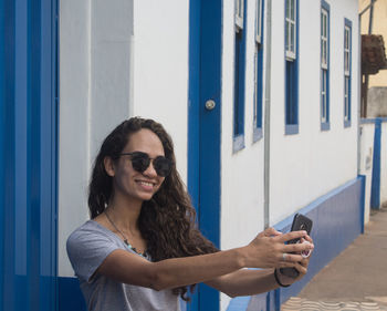 Smiling young woman taking selfie while standing by wall