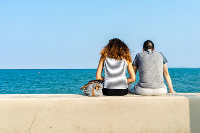 Rear view of couple sitting on retaining wall by sea against sky