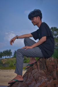 Young man sitting on rock against sky