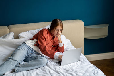 Thoughtful interested teen girl student lying on bed use laptop college study online webinar at home