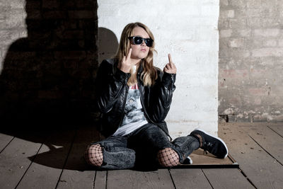 Girl showing middle finger while sitting on footpath against wall