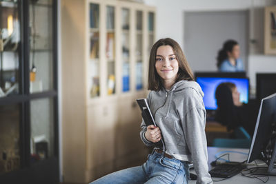 Portrait of smiling high school female student sitting with books on desk in classroom