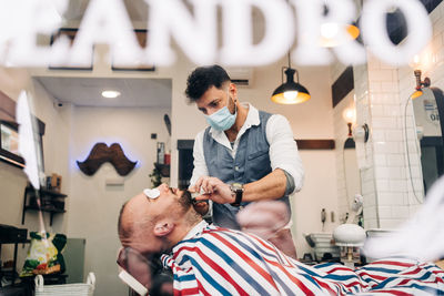 Through glass wall view of anonymous masculine stylist shaving beard of man with cotton pads on eyes using straight razor in beauty salon