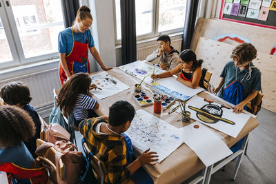 High angle view of male and female pupils painting while sitting near desk in art class at elementary school