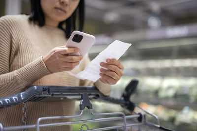 Young woman examining list while using smart phone at supermarket