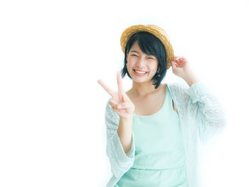 Portrait of smiling young woman showing peace sign against white background