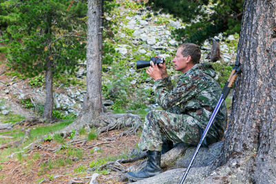Man holding binoculars while sitting against tree trunk in forest